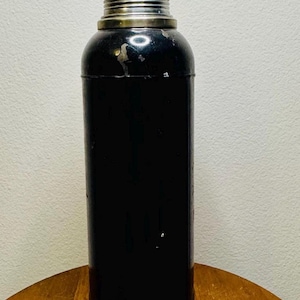 May include: A vintage black cocktail shaker with a silver-colored top and base. The cylindrical body has a glossy finish, and the shaker appears to be made of metal. The base is a gold color. The shaker is likely used for mixing drinks.