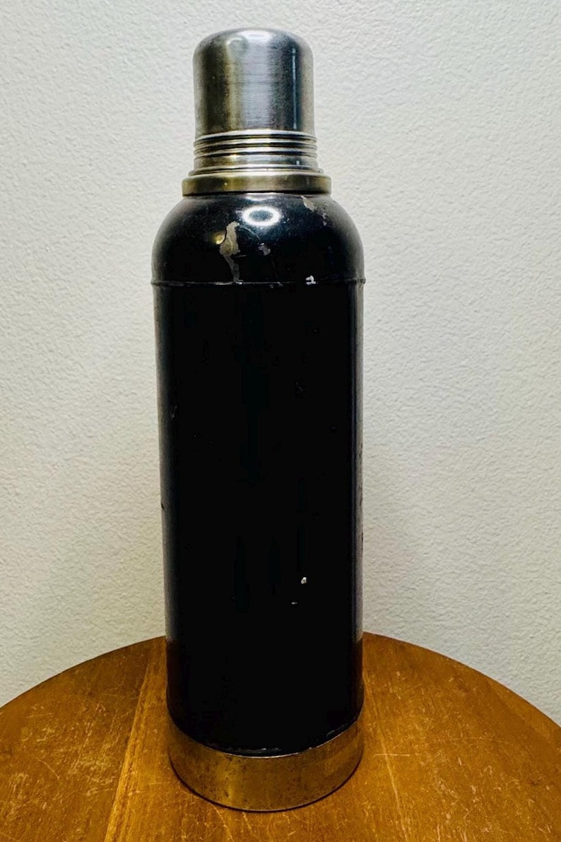 May include: A vintage black cocktail shaker with a silver-colored top and base. The cylindrical body has a glossy finish, and the shaker appears to be made of metal. The base is a gold color. The shaker is likely used for mixing drinks.