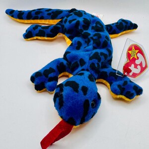 May include: A blue plush toy lizard with black spots and a red tongue. The toy has yellow accents on its underside and limbs. A red and yellow heart-shaped tag is attached.