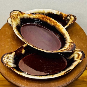 May include: Three vintage oval serving dishes. The dishes have a deep burgundy interior and a brown and tan exterior with handles. The dishes are stacked on a wooden surface.