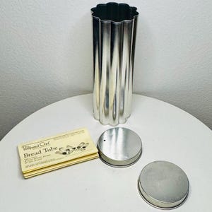May include: A silver-colored, fluted bread tube with two matching lids and a pamphlet. The bread tube has a scalloped top and bottom. The pamphlet reads "Pampered Chef Bread Tube". The items are on a white surface.