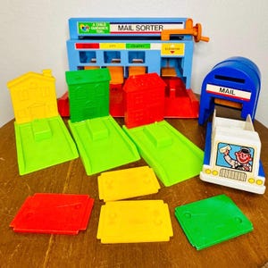 May include: A colorful vintage toy mail sorter with a blue base, orange handle, and the text "MAIL SORTER." Includes red, green, and yellow houses, ramps, and mailboxes. A white and blue toy mail truck is also present.