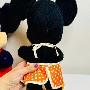 May include: A vintage Minnie Mouse plush toy, featuring black fur, large round ears, and an orange apron with white polka dots. The toy has yellow shoes and a white bow. The back of the toy is visible.