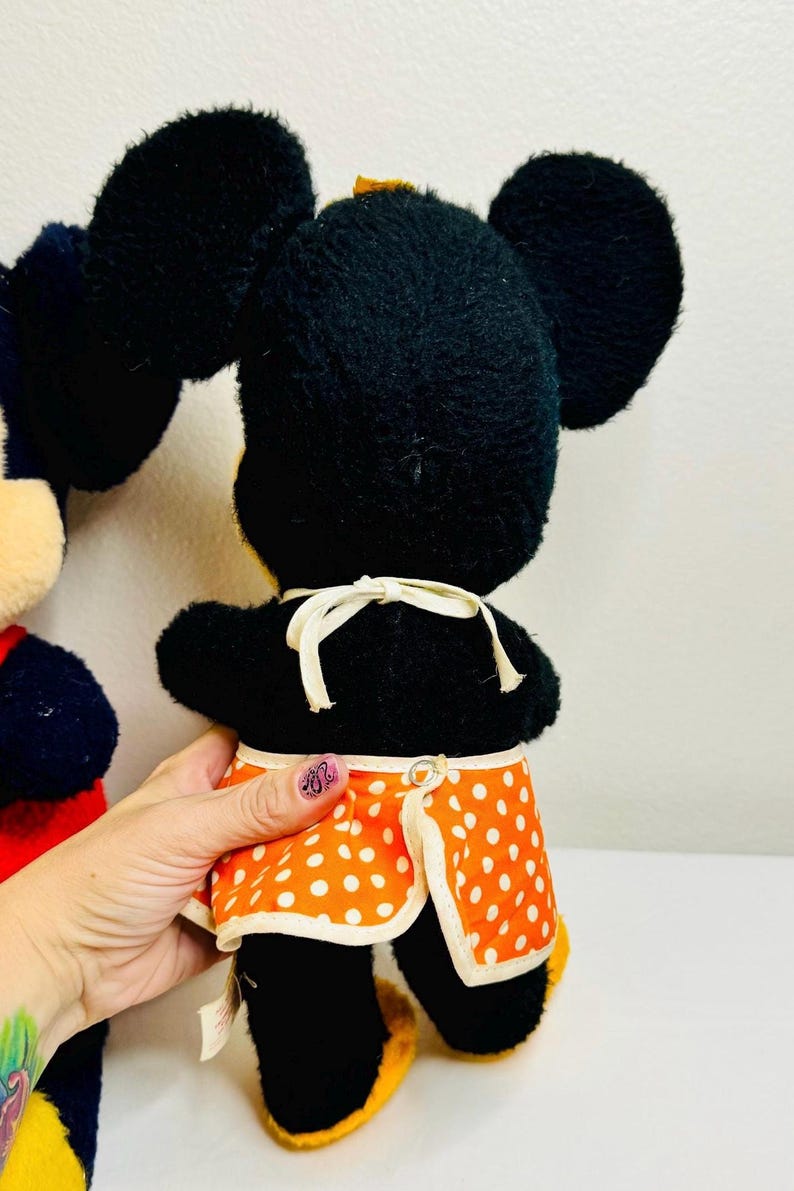 May include: A vintage Minnie Mouse plush toy, featuring black fur, large round ears, and an orange apron with white polka dots. The toy has yellow shoes and a white bow. The back of the toy is visible.