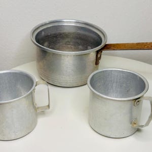 May include: A set of three aluminum camping cookware items. The set includes two mugs with handles and a small saucepan with a wooden handle. The items are silver and appear to be vintage.