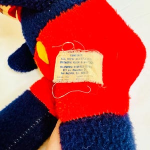 May include: A vintage stuffed toy with a red torso, navy blue limbs, and a yellow accent. The toy has a tag with text that reads "CONTENTS ALL NEW MATERIAL SYNTHETIC FOAM & COTTON CALIFORNIA STUFFED TOY".