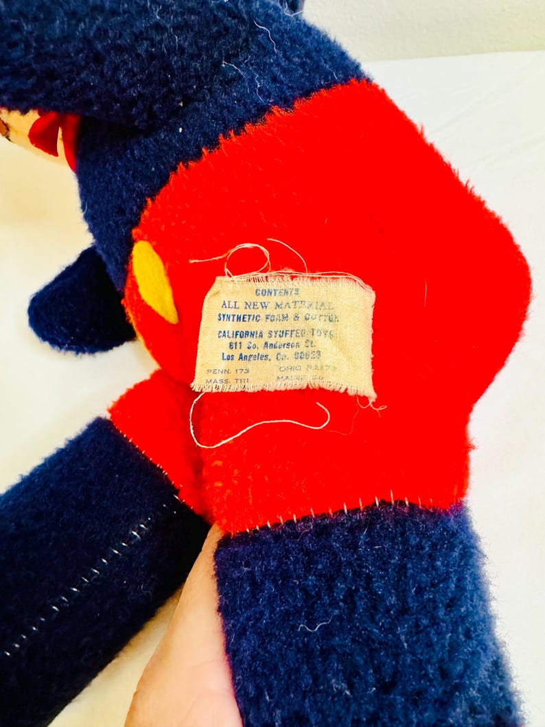 May include: A vintage stuffed toy with a red torso, navy blue limbs, and a yellow accent. The toy has a tag with text that reads "CONTENTS ALL NEW MATERIAL SYNTHETIC FOAM & COTTON CALIFORNIA STUFFED TOY".