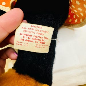 May include: A close-up of a stuffed toy with a tag. The tag reads "CONTENTS ALL NEW MATERIAL SYNTHETIC FIBERS CALIFORNIA STUFFED TOYS 611 So. Anderson St. Los Angeles, Ca. 90023". The toy has black, orange, and tan sections.