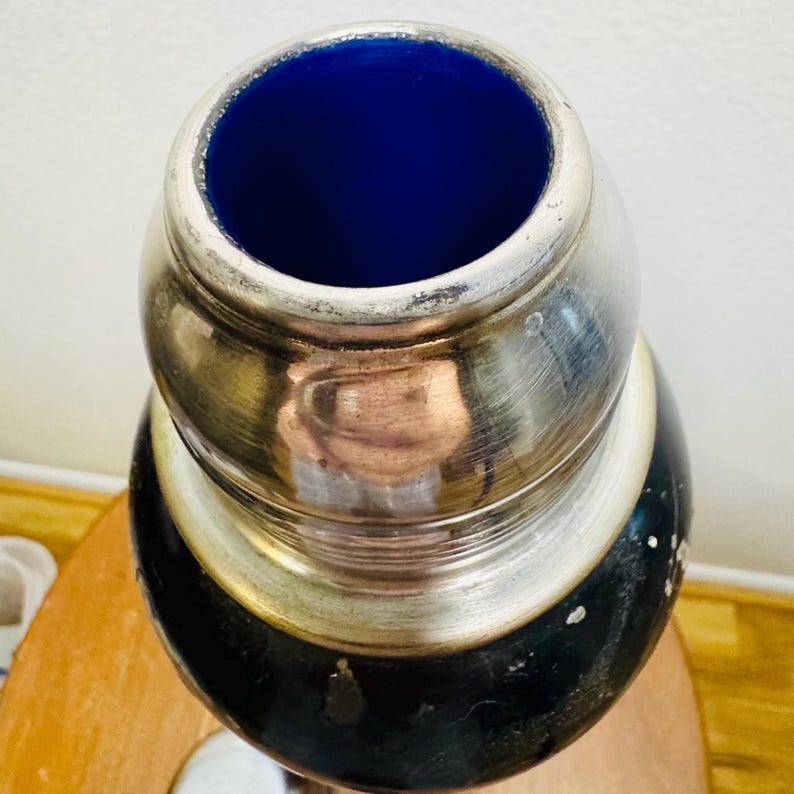 May include: A silver-colored gourd-shaped vessel with a dark blue interior. The vessel has a silver rim and a black base with white speckles. The gourd is sitting on a wooden surface.