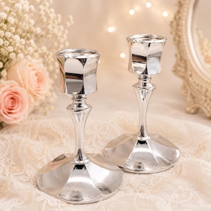 May include: A pair of silver-toned metal candle holders featuring a faceted, hexagonal base and a matching hexagonal top cup. The stems are slender and tapered, connecting the wide base to the candle cup in a polished, reflective finish.