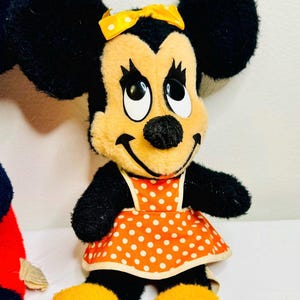 May include: A plush Minnie Mouse doll with black ears, arms, and a smiling face. She wears an orange dress with white polka dots and a yellow bow. Her feet are yellow.