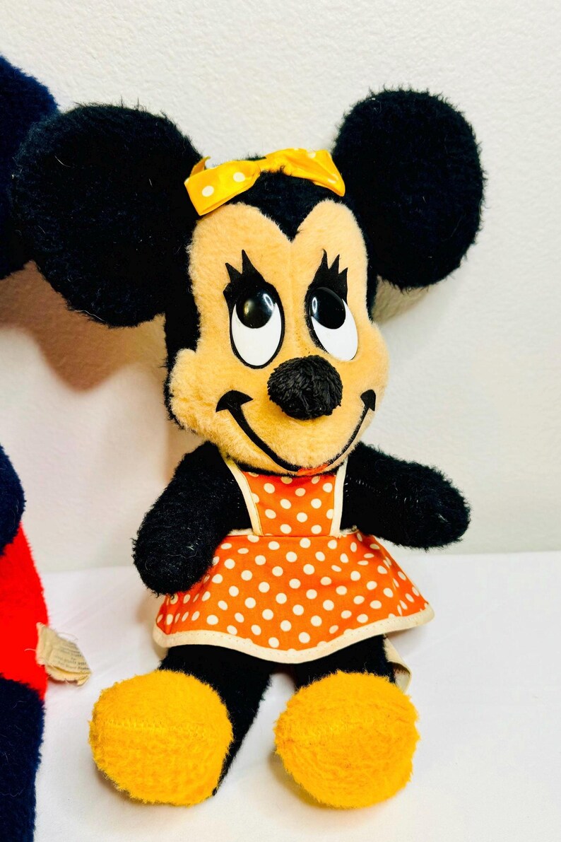 May include: A plush Minnie Mouse doll with black ears, arms, and a smiling face. She wears an orange dress with white polka dots and a yellow bow. Her feet are yellow.