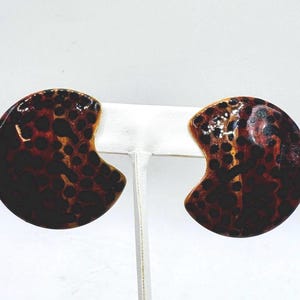 May include: A pair of large, abstract earrings with a tortoiseshell pattern. The earrings are a half-moon shape with a dark brown and amber color scheme. The earrings are displayed on a white earring stand.