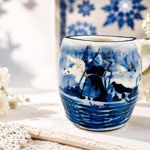 May include: A ceramic mug featuring a hand-painted blue and white scene of a windmill near water. The mug has a rounded body, a thick handle, and a glossy finish. The design includes dark blue brushstrokes depicting the windmill structure.