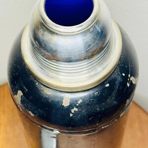 May include: Vintage cocktail shaker with a black body and a silver-colored top. The interior of the shaker's top is a vibrant blue. The shaker shows signs of wear, adding to its antique charm. A classic barware item.