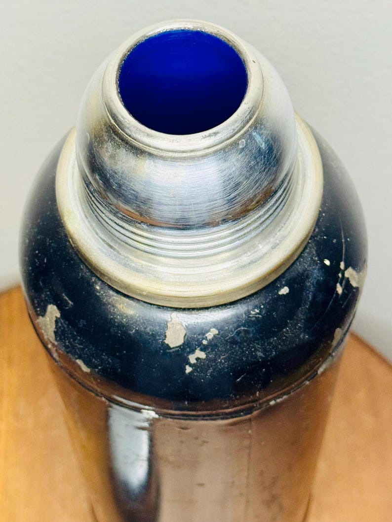 May include: Vintage cocktail shaker with a black body and a silver-colored top. The interior of the shaker's top is a vibrant blue. The shaker shows signs of wear, adding to its antique charm. A classic barware item.