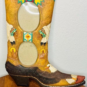 May include: A decorative, western-themed photo frame shaped like a cowboy boot. The frame is tan with brown and black accents, featuring Native American figures, an eagle, and two oval openings for photos. The boot is approximately 12 inches tall.