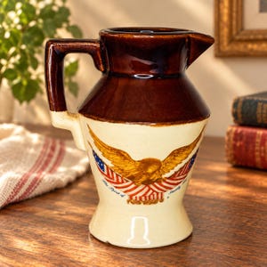May include: A ceramic pitcher with a dark brown glazed top and a cream-colored base. The front features a graphic of a golden eagle with outstretched wings positioned above two crossed American flags and the text The Spirit of Seventy Six.
