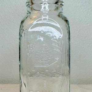 May include: Clear glass jar with a cork stopper. The jar is rectangular with rounded corners and embossed with the words "1 POUND PURE HONEY". The jar has a decorative design of a beehive and flowers.