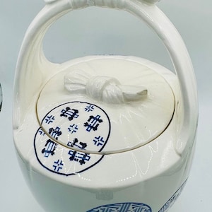 May include: White ceramic teapot with a blue and white design. The teapot has a handle shaped like a bow and a lid with a knob shaped like a bow. The teapot is decorated with Chinese characters.