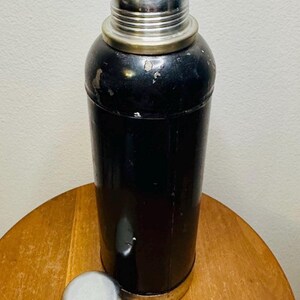 May include: A vintage black cocktail shaker with a silver top and a black stopper. The shaker is cylindrical with a silver base and a separate silver cap. The cap is placed next to the shaker on a wooden surface.