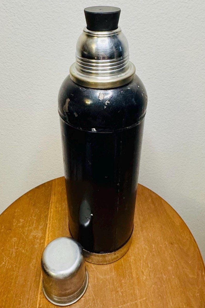 May include: A vintage black cocktail shaker with a silver top and a black stopper. The shaker is cylindrical with a silver base and a separate silver cap. The cap is placed next to the shaker on a wooden surface.