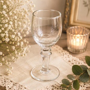 May include: A clear glass goblet featuring a rounded bowl, a thick stem with a decorative ringed detail, and a wide circular base. The glass is transparent and smooth, designed with a classic silhouette that tapers from the rim to the stem.