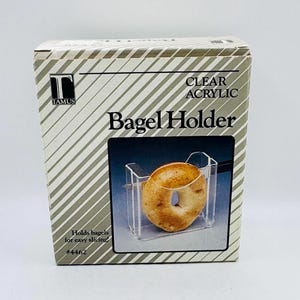 May include: A rectangular cardboard product box for a Tamus clear acrylic bagel holder. The box features diagonal grey and white stripes, the text "CLEAR ACRYLIC Bagel Holder", "Holds bagels for easy slicing!", and "#4462" with a product photo.