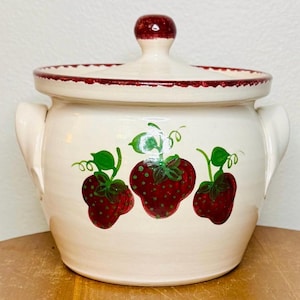 May include: Cream-colored ceramic jar with a lid, featuring a hand-painted strawberry design. The jar has a red rim and handle, and a red knob on the lid. The strawberries are red with green leaves.