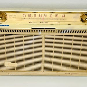 May include: Vintage General Electric radio with a cream-colored casing and a speaker grill. The radio features a tuning dial with numbers and the words "PHONO RADIO". The radio has two gold knobs and the text "PULL ON".