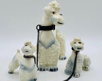 May include: Three ceramic poodle figurines in cream and blue. The largest poodle stands upright, while the other two are in a sitting and standing position. Each poodle has a black collar and leash. The figurines have a vintage aesthetic.