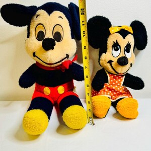 May include: Two plush Mickey and Minnie Mouse dolls. Mickey is wearing red shorts with yellow buttons, a red bow tie, and yellow shoes. Minnie is wearing a polka dot dress and a yellow bow. Mickey is approximately 15 inches tall.