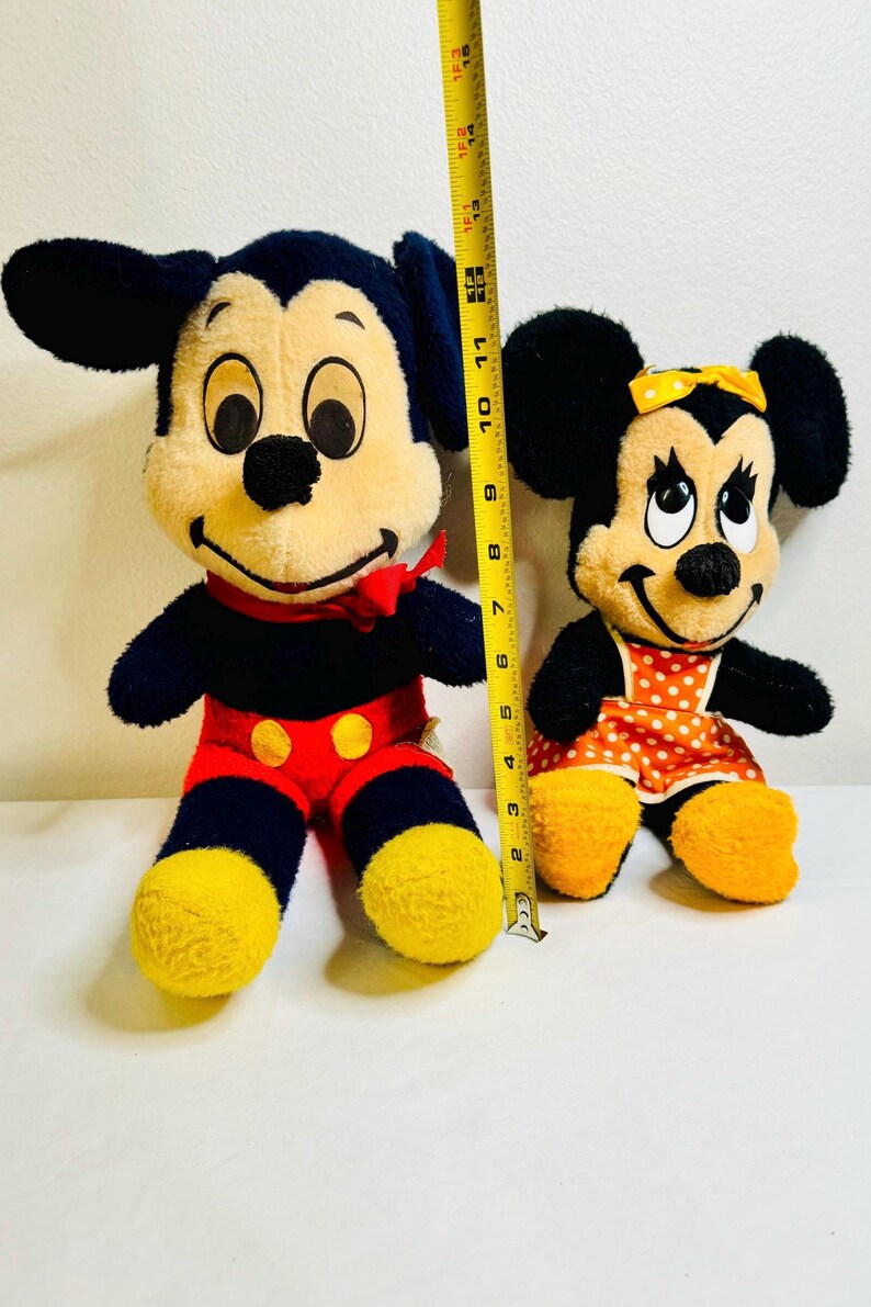 May include: Two plush Mickey and Minnie Mouse dolls. Mickey is wearing red shorts with yellow buttons, a red bow tie, and yellow shoes. Minnie is wearing a polka dot dress and a yellow bow. Mickey is approximately 15 inches tall.