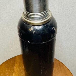 May include: A vintage cocktail shaker with a black body and a silver-colored top. The shaker has a cylindrical shape with a rounded top and a silver band around the middle. The surface shows signs of wear, suggesting it's an antique barware item.
