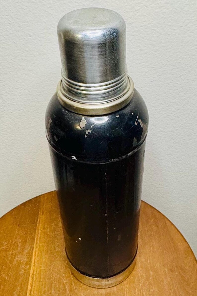 May include: A vintage cocktail shaker with a black body and a silver-colored top. The shaker has a cylindrical shape with a rounded top and a silver band around the middle. The surface shows signs of wear, suggesting it's an antique barware item.