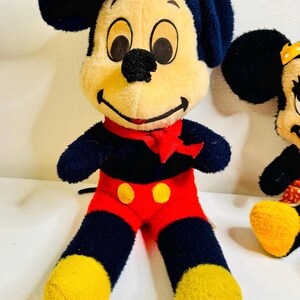 May include: A plush Mickey Mouse doll with a black body, red shorts, and yellow feet. The doll has a cream-colored face, black eyes, and a red bow tie. Another similar doll is partially visible.