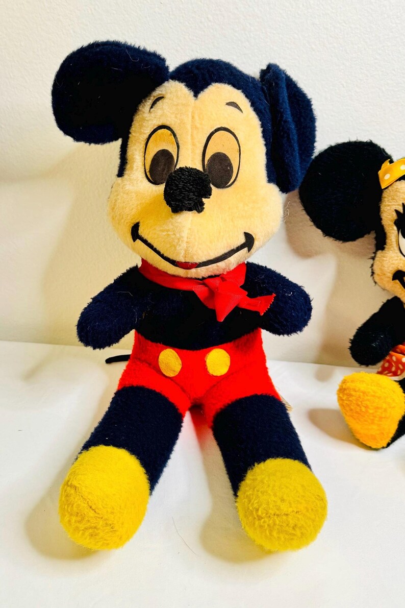 May include: A plush Mickey Mouse doll with a black body, red shorts, and yellow feet. The doll has a cream-colored face, black eyes, and a red bow tie. Another similar doll is partially visible.