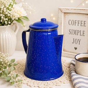 May include: A vibrant cobalt blue enamel coffee percolator with a speckled white finish, featuring a matching lid, a sturdy side handle, and a wide pouring spout. The coffee pot has a smooth, glossy surface and a classic tapered cylindrical shape.