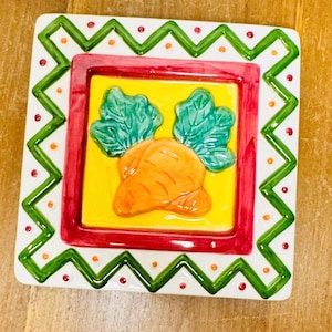 May include: Square ceramic plate with a carrot design. The plate features a yellow center with an orange carrot and green leaves, framed by a red border. The outer edge has a white background with green zigzags and red and orange dots.