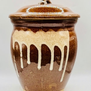 May include: A brown ceramic jar with a lid and a white glaze design. The jar has a rounded body and a slightly flared opening. The lid has a small handle.