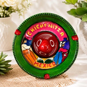 May include: A round ceramic ashtray with a green border and a central red sombrero shape. The design features the text CHICHEN ITZA and MEXICO in yellow letters, accompanied by painted figures in orange and blue clothing on a pink background.