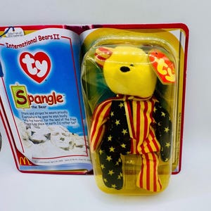May include: A yellow and red beanie baby bear wearing a red, white, and blue outfit with stars and stripes. The bear is packaged in a clear plastic bag with a Ty tag and a McDonald's logo. The packaging also includes the text "International Bears II" and "Spangle the Bear".