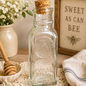 May include: A clear glass bottle with a cork stopper, featuring an embossed beehive design and the text 1 POUND PURE HONEY. The square-shaped bottle has a narrow neck and a wide base, showcasing a classic design for storing liquid honey.