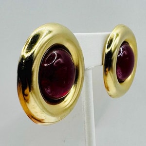 May include: A pair of gold-toned clip-on earrings. Each earring features a large, round, deep red center stone set within a thick, circular gold frame. The earrings are displayed on a white stand against a white background.