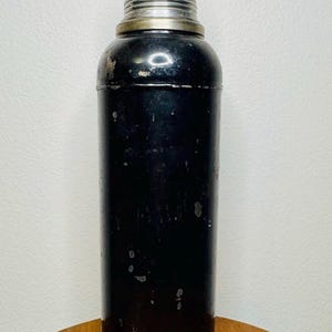 May include: Vintage cocktail shaker with a black body and a silver-colored top and base. The shaker has a cylindrical shape with a rounded top and a cap. The metal shows signs of wear, adding to its vintage appeal. A classic barware item.