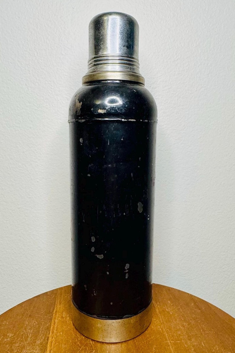 May include: Vintage cocktail shaker with a black body and a silver-colored top and base. The shaker has a cylindrical shape with a rounded top and a cap. The metal shows signs of wear, adding to its vintage appeal. A classic barware item.