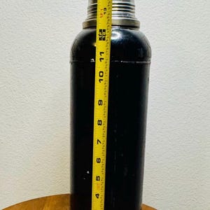 May include: A black cocktail shaker with a silver top and base, measuring approximately 15 inches tall. The shaker is cylindrical with a slightly rounded top and bottom, and is being measured with a yellow tape measure.