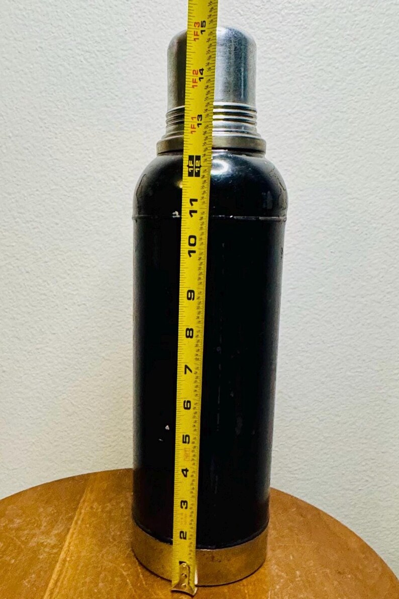 May include: A black cocktail shaker with a silver top and base, measuring approximately 15 inches tall. The shaker is cylindrical with a slightly rounded top and bottom, and is being measured with a yellow tape measure.