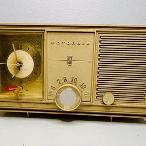 May include: Vintage Motorola radio and clock combo in a cream-colored case. The radio features a clock with a gold face and black numerals, a tuning dial, and a speaker grill. The clock shows the time as 12:00.