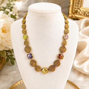May include: A gold-toned necklace featuring a series of circular, textured metal links resembling coiled rope. Interspersed among the gold links are five faceted, round gemstones in vibrant shades of lime green, purple, amber, yellow, and pink.